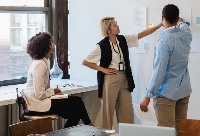 Two men and a woman in an office look at a whiteboard. 