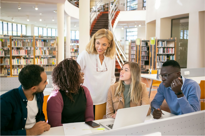 Five people in a library around a laptop. One woman in the middle is standing up.