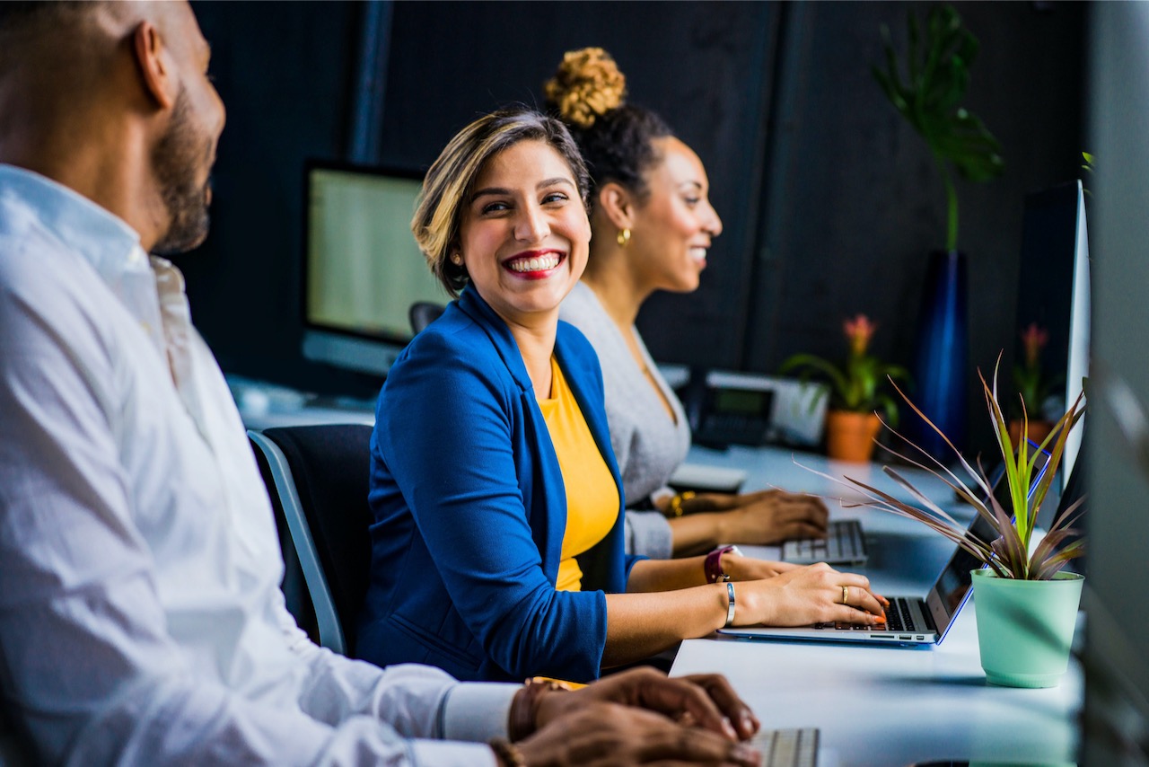 Two women sitting at their own laptops. The woman in the middle is smiling towards a man who is closer to the camera. 